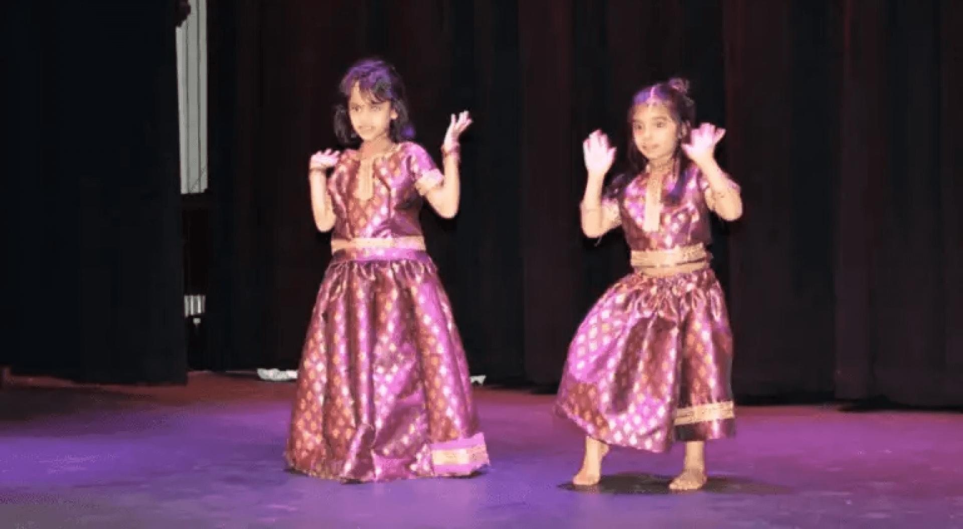 Two young girls dressed in traditional Indian dance costumes perform on stage, their expressions and hand gestures showing concentration and joy during a kids dance performance.