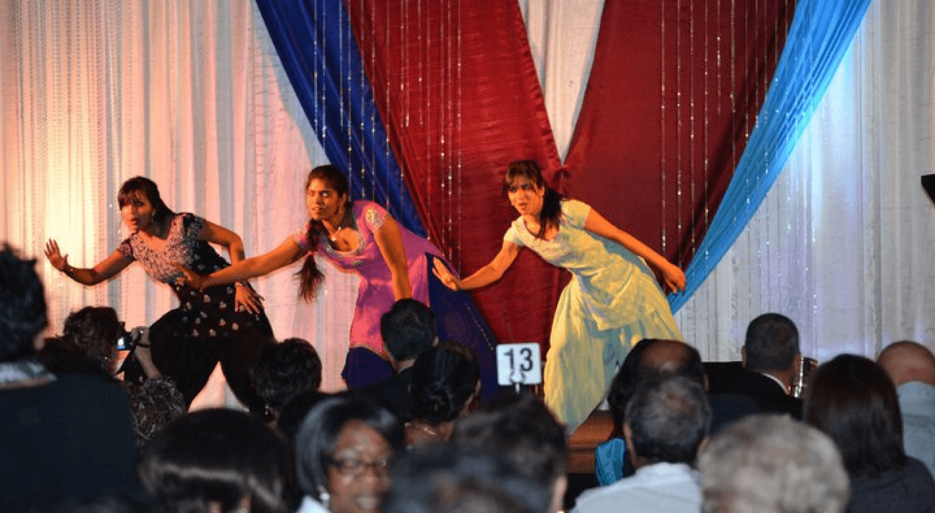 Three dancers performing on a stage decorated with red, white, and blue drapes, in front of an audience. The dancers are captured mid-movement, showcasing their expressive dance styles at what appears to be a community event or celebration.