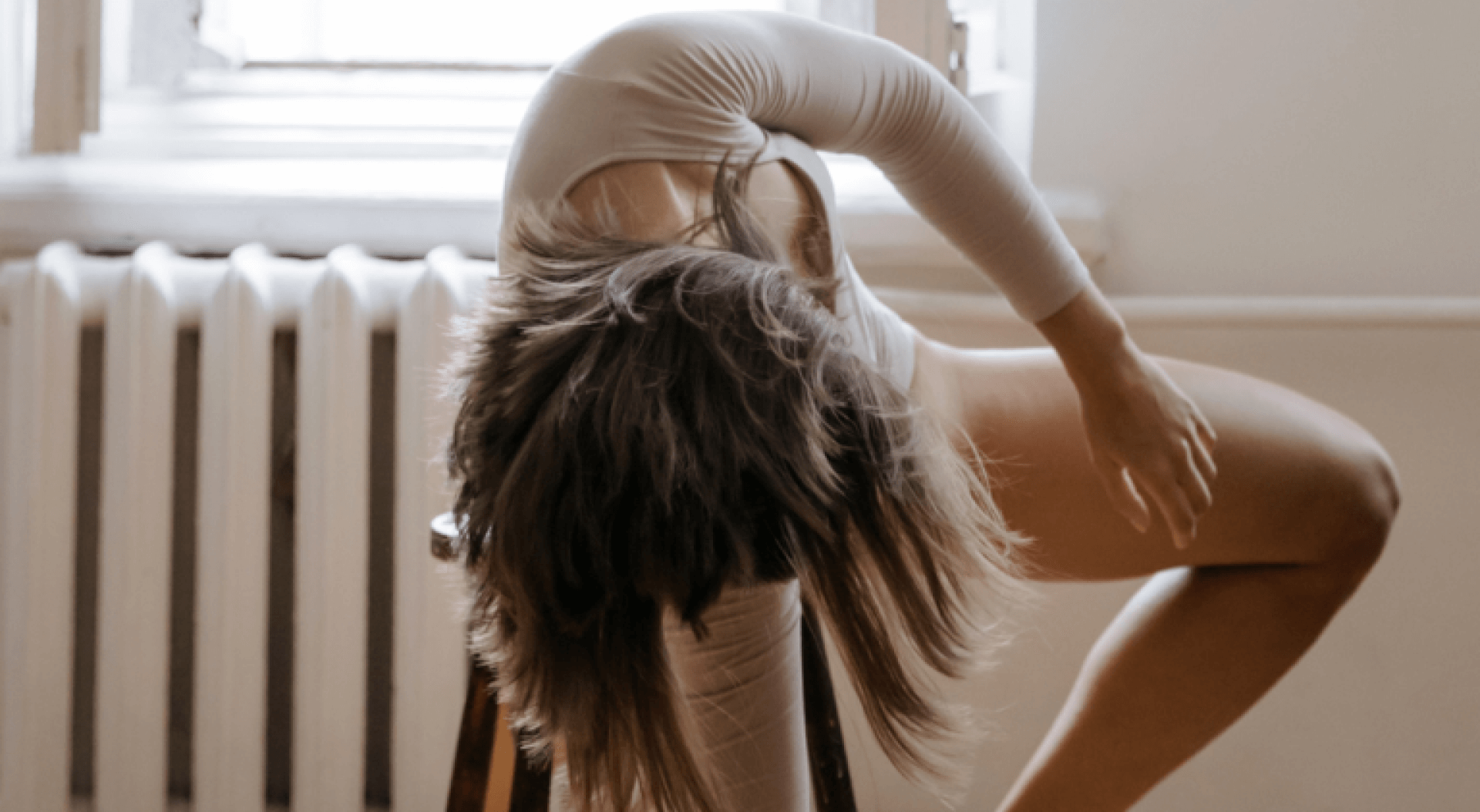 A dancer in a neutral-toned bodysuit performs an intricate pose on a wooden chair, demonstrating flexibility and strength in a bright room with a vintage radiator and window providing natural light. Her movement and form are the focus of this artistic dance practice setting.