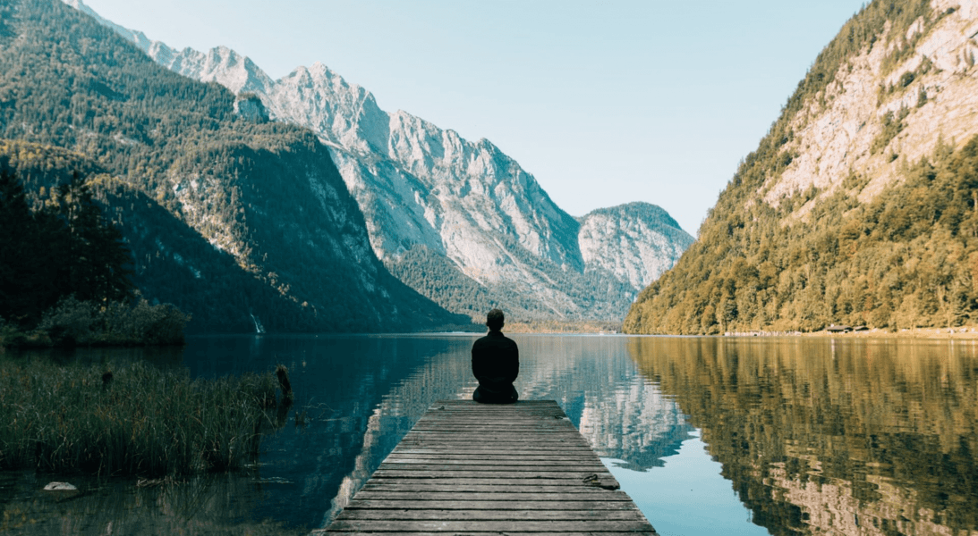 A person sitting peacefully at the end of a wooden dock, overlooking a serene mountain lake with reflections of the surrounding forested hills on the water's surface, under a clear sky, creating a tranquil and picturesque natural setting.