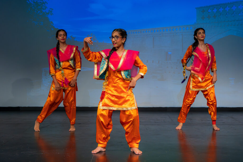 Three dancers in bright orange and pink traditional Indian costumes perform a lively dance on stage, with a backdrop of an architectural scene, one dancer in the foreground energetically leading the group.