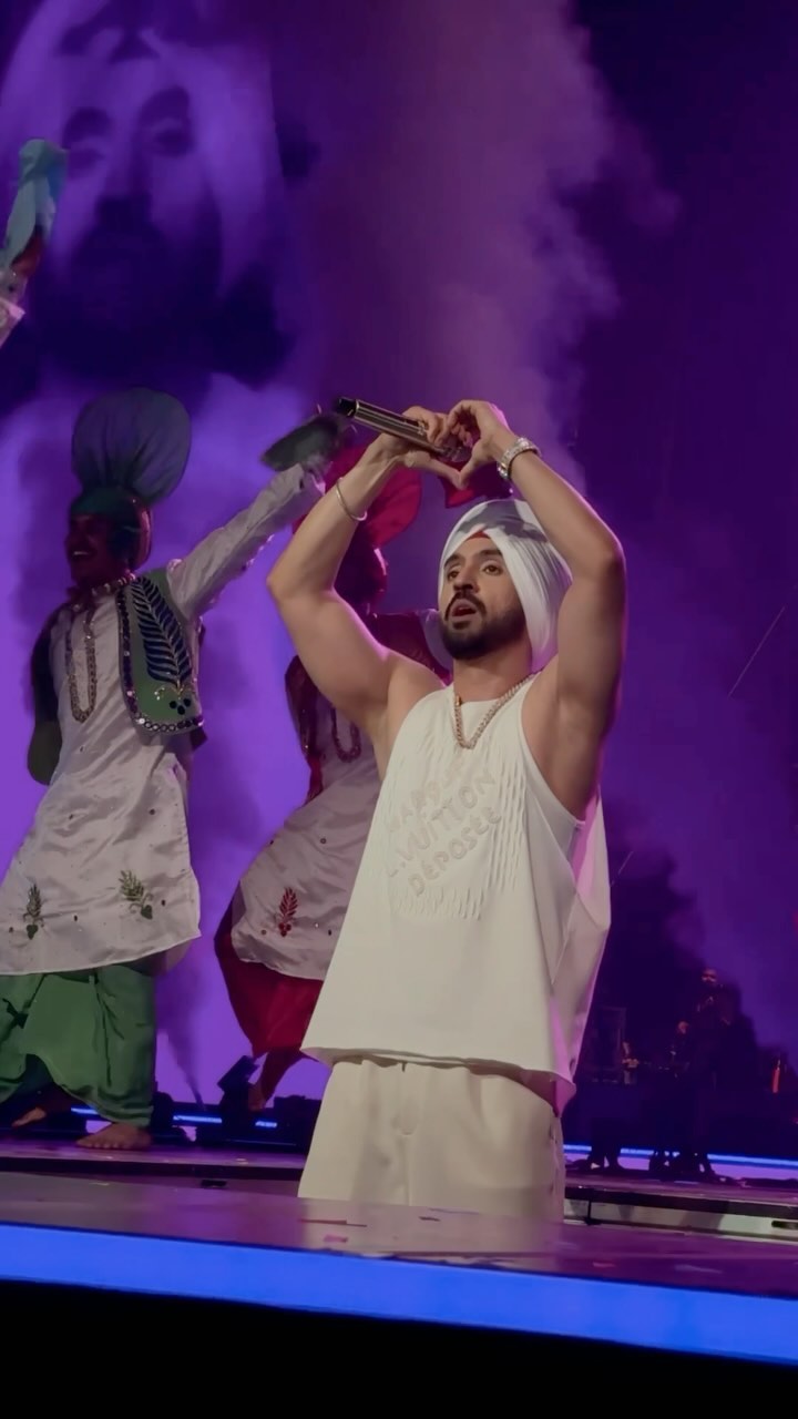 Diljit Dosanjh wearing a white turban and sleeveless white outfit makes a heart shape with his hands on stage, with vibrant dancers in traditional attire in the background and colorful stage lights creating a dynamic atmosphere.